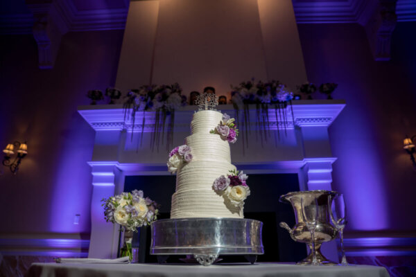 Beautiful white wedding cake with flower accents in front of mantel with purple uplights